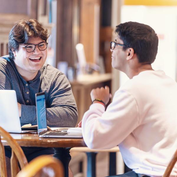 Two male student sit talking at a table. On the table is a laptop with several stickers on the back.